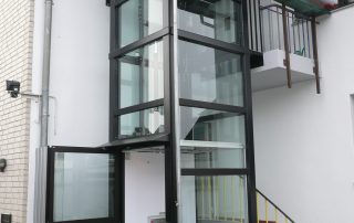 Außenaufzug aus Glas neben Eingang aber Zugang über Balkonkonstruktion