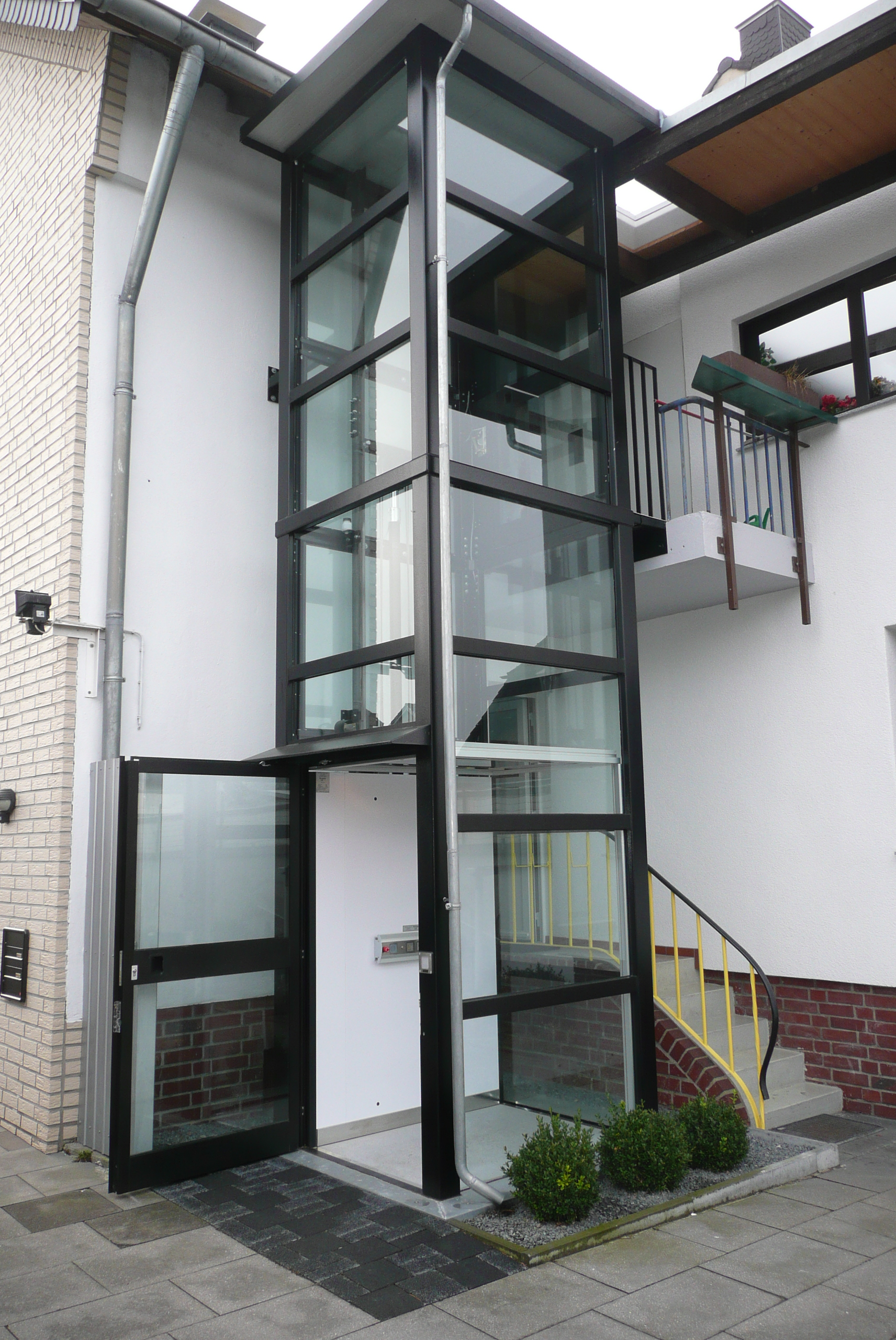 Außenaufzug aus Glas neben Eingang aber Zugang über Balkonkonstruktion